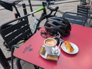 coffee, cake, bicycle helmet, glasses, and gloves on a table with bicycle leaning against it