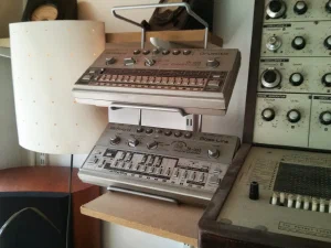 A shelf-mounted vintage synthesizer setup featuring an EMS VCS3 analog synthesizer in a wooden case with patch matrix and joystick, alongside a Roland TR-606 drum machine and Roland TB-303 Bass Line, with a small lamp and decorative items visible.