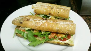 A sandwich on a white plate, made with a seeded baguette, filled with arugula, tomato, bacon, and cheese