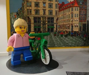 A large LEGO minifigure standing next to a green LEGO bicycle, in front of a LEGO mosaic of a city street scene