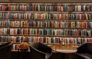 A tall wall of bookshelves packed with colorful paperbacks, fronted by leather armchairs, a round wooden table, and an antique globe
