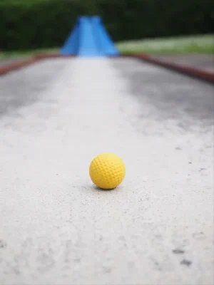 A single yellow dimpled mini-golf ball resting on a worn concrete putting lane, with a blue ramp obstacle blurred in the background in front of a green hedge