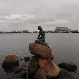 The Little Mermaid statue on a rock in Copenhagen harbour, with the industrial harbour skyline behind it