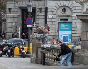 A skateboarder popping a trick off a stone ledge at Plaça de Catalunya in Barcelona while a filmer crouches to capture it