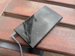 A black Sony Walkman music player lying on a wooden bench, screen showing a track playing, with a grey headphone cable plugged in