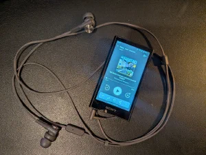 A black Sony Walkman music player on a dark leather surface, screen showing a podcast episode of The Europeans ready to play, with grey Sony in-ear headphones coiled around it
